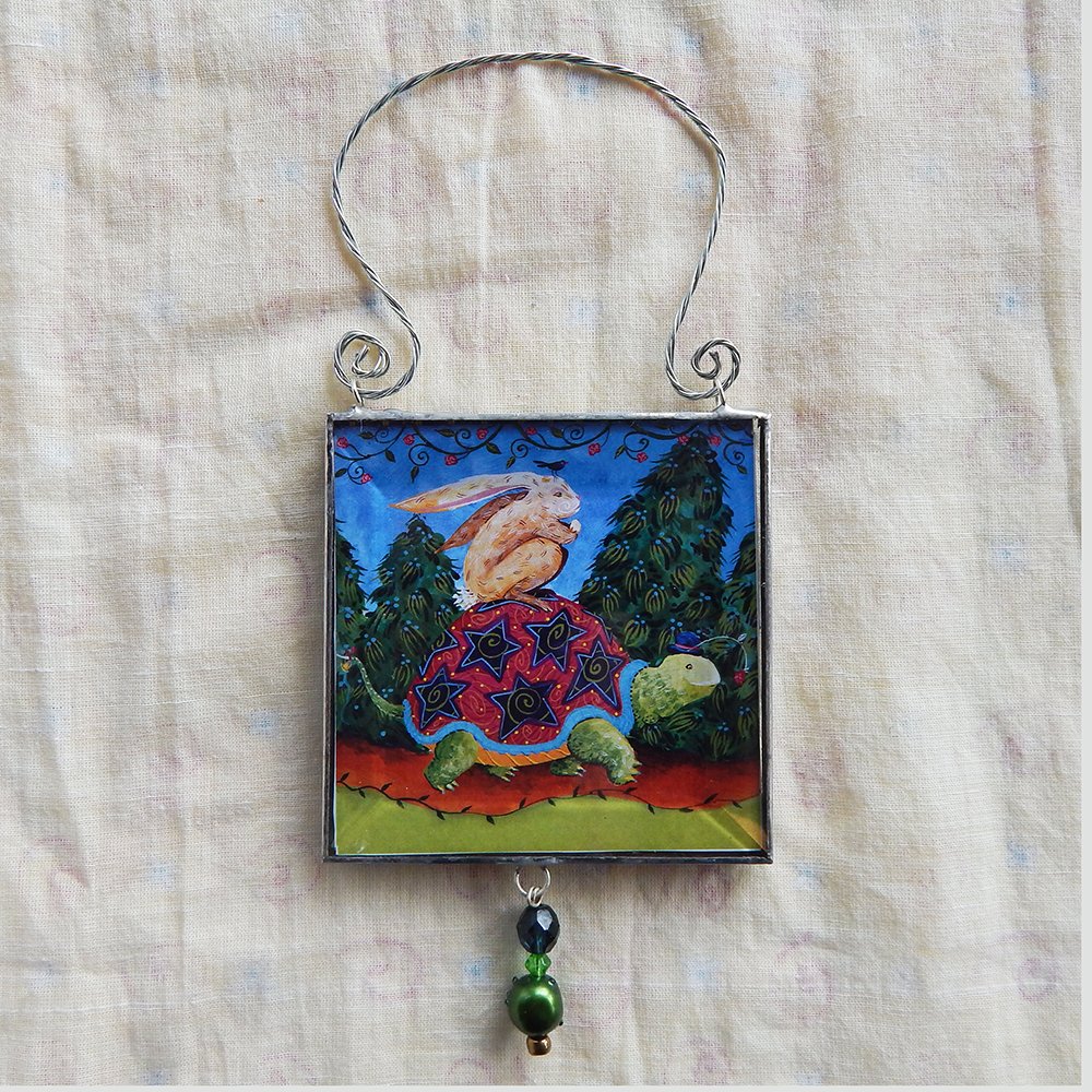 Danasimson.com The Journey is the thing, double sided beveled glass ornament with a hare riding on a tortoise image. Twisted wire handle and bead detail.